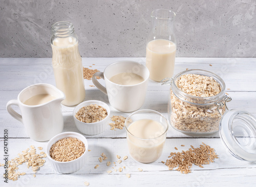 Set of different containers with oat milk, oat seeds and flakes on white wooden table on concrete background. 