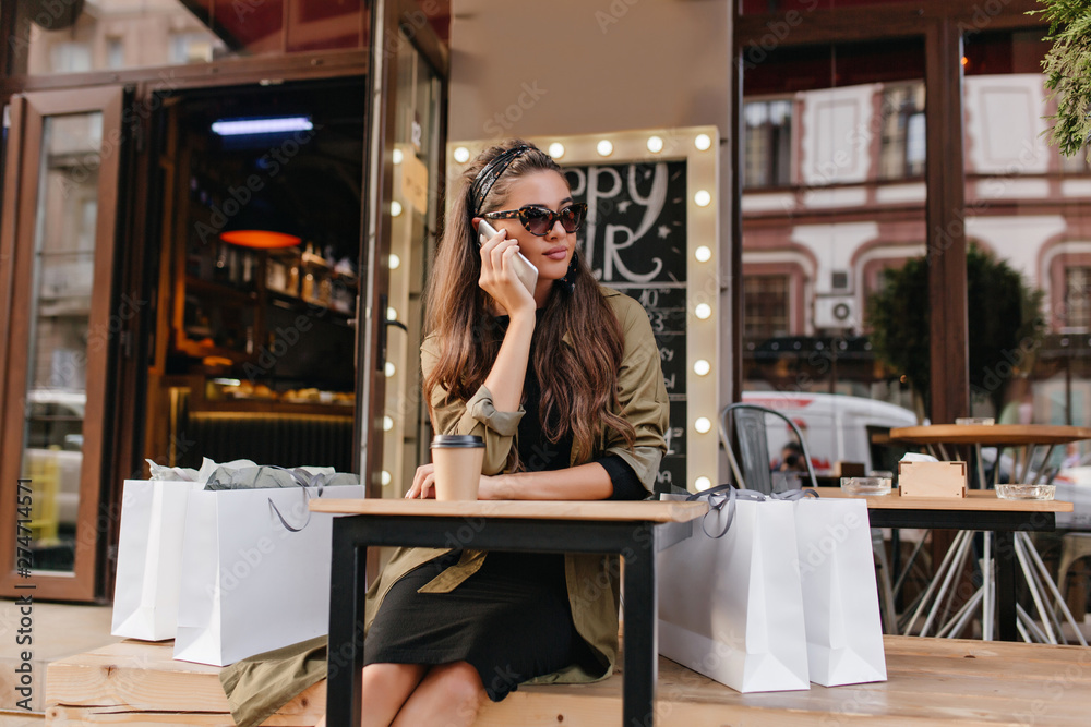 Bored girl calling someone while sitting in outdoor cafe after shopping. Long-haired female model in stylish autumn attire talking on phone, surrounded by purchases.