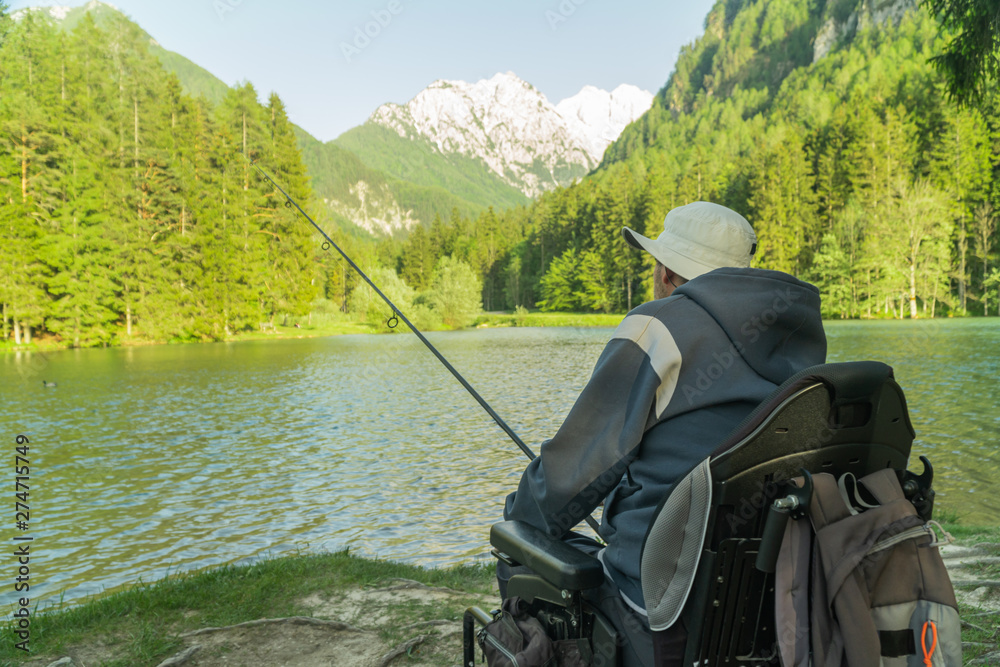 young man in a wheelchair fishing at the beautiful lake on a sunny day, with mountains in the back young man in a wheelchair fishing at the beautiful lake on a sunny day, with mountains in the back