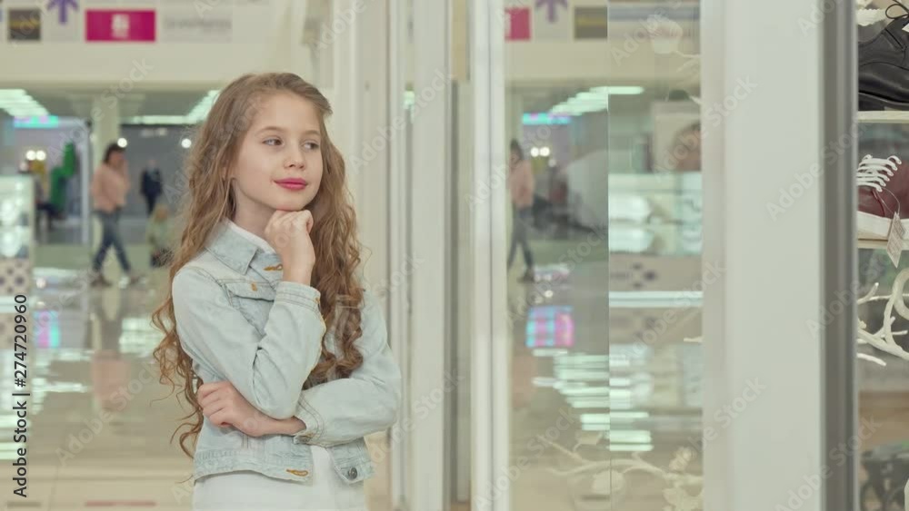 Cute little girl smiling to the camera, examining fashion store display. Adorable beautiful girl looking at clothing boutique showcase. Happy lovely girl at the shopping mall Cute little girl smiling to the camera, examining fashion store display. Adorable beautiful girl looking at clothing boutique showcase. Happy lovely girl at the shopping mall