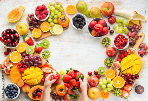 Raw healthy breakfast frame, cut fruits, strawberries raspberries oranges plums apples kiwis grapes blueberries mango on white table, child's hands holding food, copy space, top view, selective focus