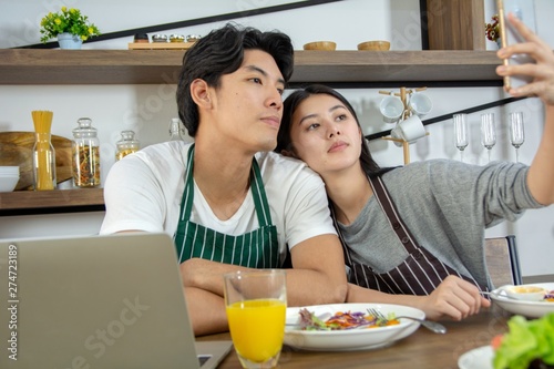 Portrait of cute happy couple in aporn in selfie action posting with cell phone decorated with salad and orange juice in modern kitchen