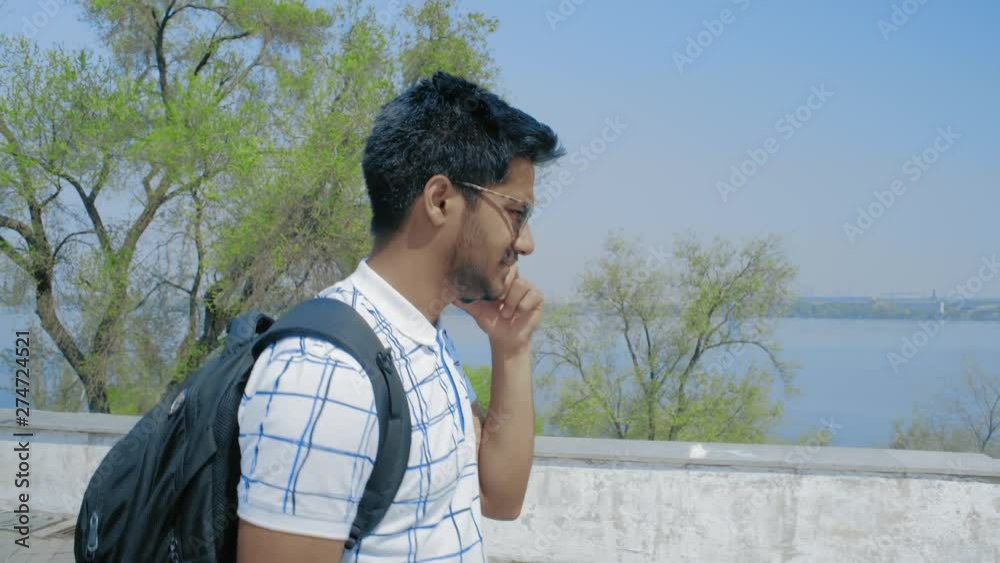 Indian Guy with Glasses and backpack Talking on the Phone, Walking in the Park against the River