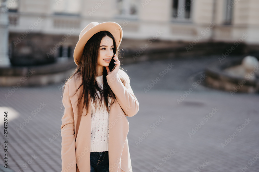 Beautiful Caucasian woman talking by phone with friend while standing on sunny street. Brunette wearing beige coat, looking away, has important conversation. Communication concept.
