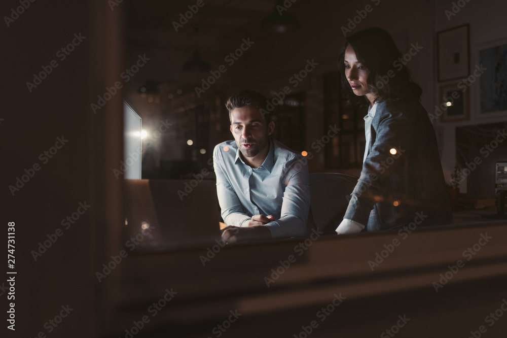 Two smiling businesspeople working on a computer together at night Two smiling businesspeople working on a computer together at night