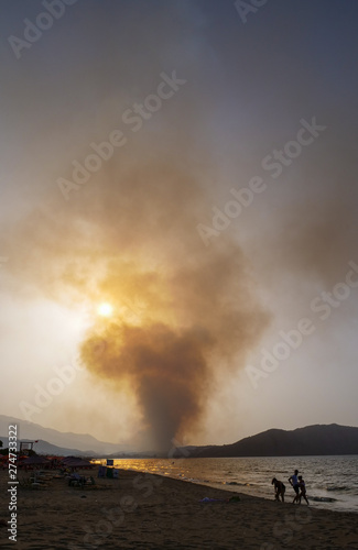 Smoke from a large fire billows into the sky as the sun sets, beach