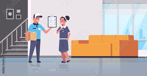 postman in uniform holding cardboard parcel box courier showing clipboard to woman recipient express delivery service concept modern living room interior flat full length