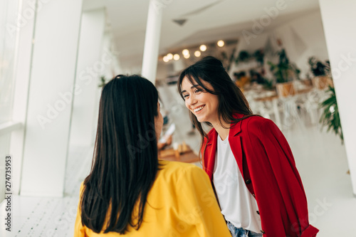 Cheerful attractive young woman in red jacket look at model in yellow and smile. Standing in white cafe together. Brunettes spend time in room. © Anton