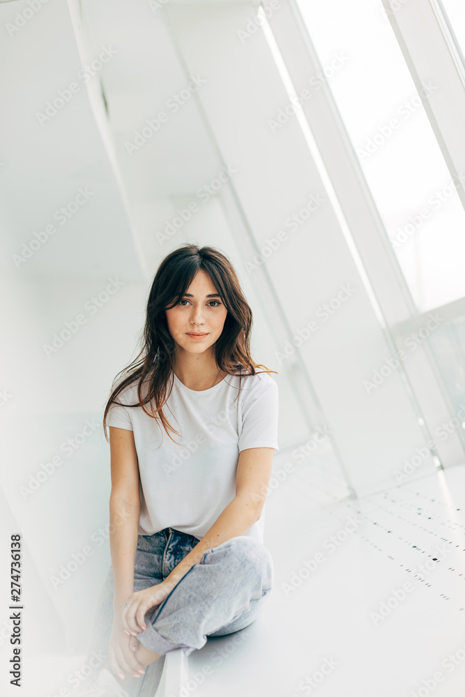 Young beautiful brunette posing on camera. Sit at window and look straight. Woman wear casual clothes. Alone in room or cafe.
