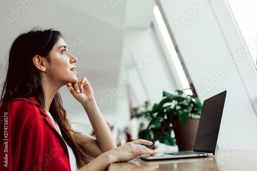 Calm, peaceful and thoughtful young buisnesswoman in red jacket working alone. Sitting at table in coworking space. Look up. Alone in white room. photo