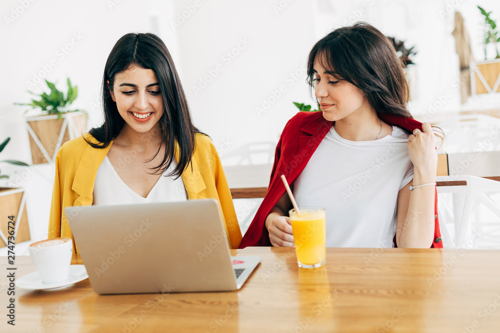Beautiful attractive young women in coworking space. Working together. Stylish businesswomen in cafe. Model on left typing on keyboard. Woman on right drink orange juice. Sit on table. Beautiful attractive young women in coworking space. Working together. Stylish businesswomen in cafe. Model on left typing on keyboard. Woman on right drink orange juice. Sit on table.