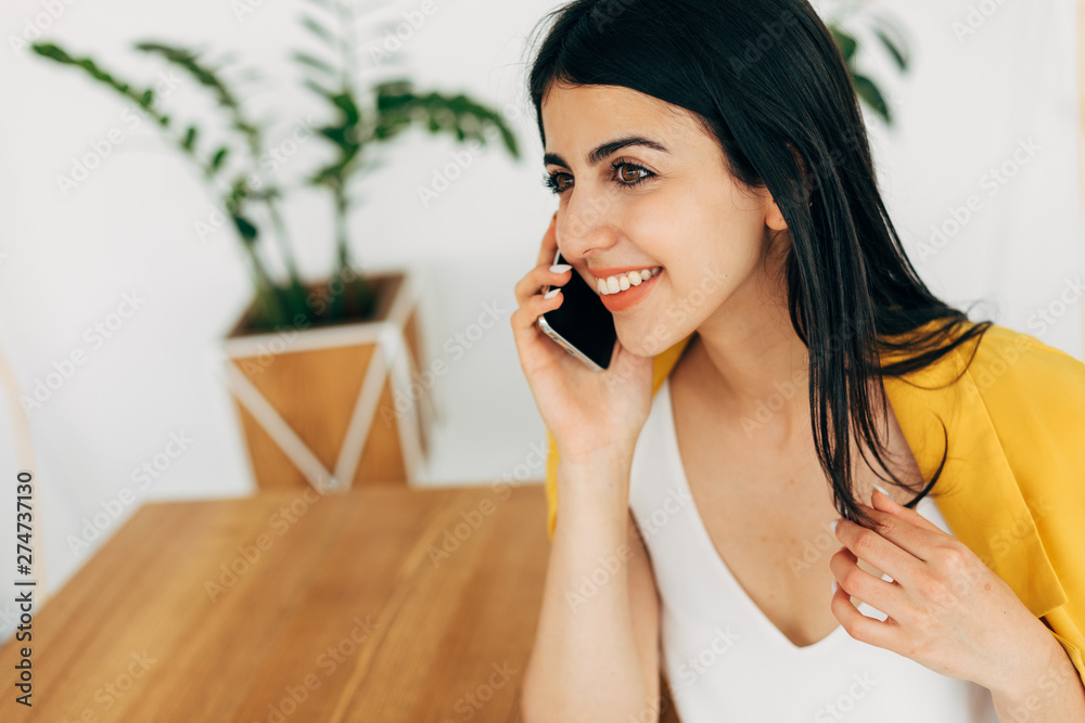 Cheerful attractive nice young woman talking on phone and smile. Sit in white room at table. Alone in coworking space. Stylish geogrian model wear yellwo jacket. Cheerful attractive nice young woman talking on phone and smile. Sit in white room at table. Alone in coworking space. Stylish geogrian model wear yellwo jacket.