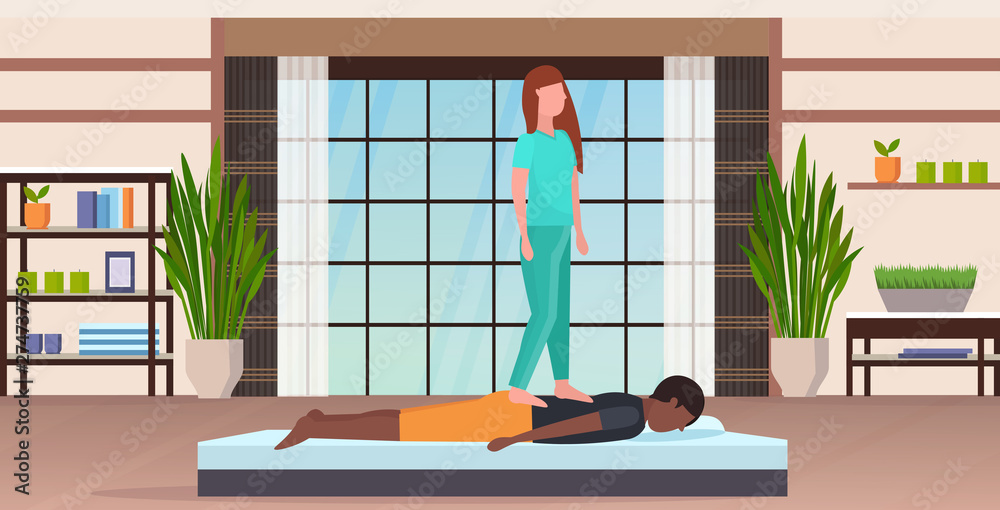 masseuse in uniform standing on patient's back doing healing treatment african american guy having massage manual therapy concept modern spa salon studio interior full length horizontal