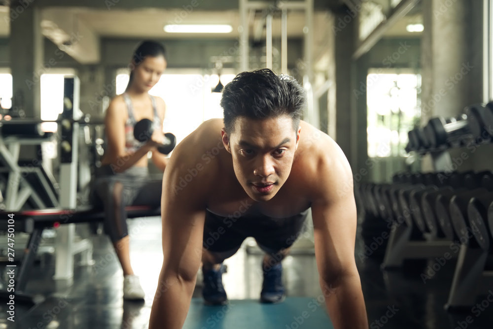 Working out at GYM concept. Young Asian handsome man working out his chest by pushing up in the gym. Young girl lift up the dumbbells in background. Health and fitness concept. Working out at GYM concept. Young Asian handsome man working out his chest by pushing up in the gym. Young girl lift up the dumbbells in background. Health and fitness concept.