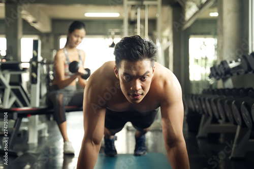 Fototapeta Naklejka Na Ścianę i Meble -  Working out at GYM concept. Young Asian handsome man working out his chest by pushing up in the gym. Young girl lift up the dumbbells in background. Health and fitness concept.