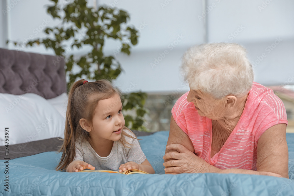 Cute girl and her grandmother reading book on bed at home Cute girl and her grandmother reading book on bed at home