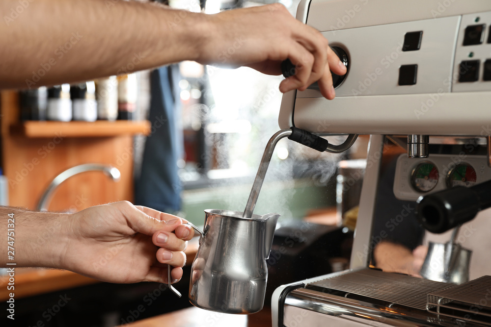Barista steaming milk in metal jug with coffee machine wand at bar counter, closeup