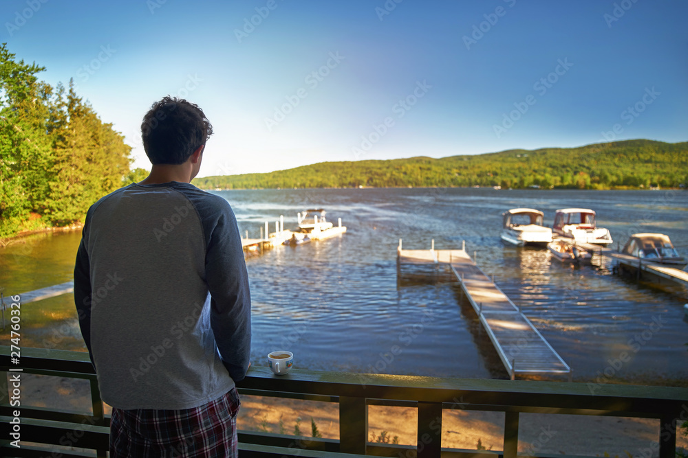 Young man stands alone on a veranda at home and looking at the lake. Early morning, sunny day. Early cold morning in the fall. Peaceful atmosphere in nature. Back view.