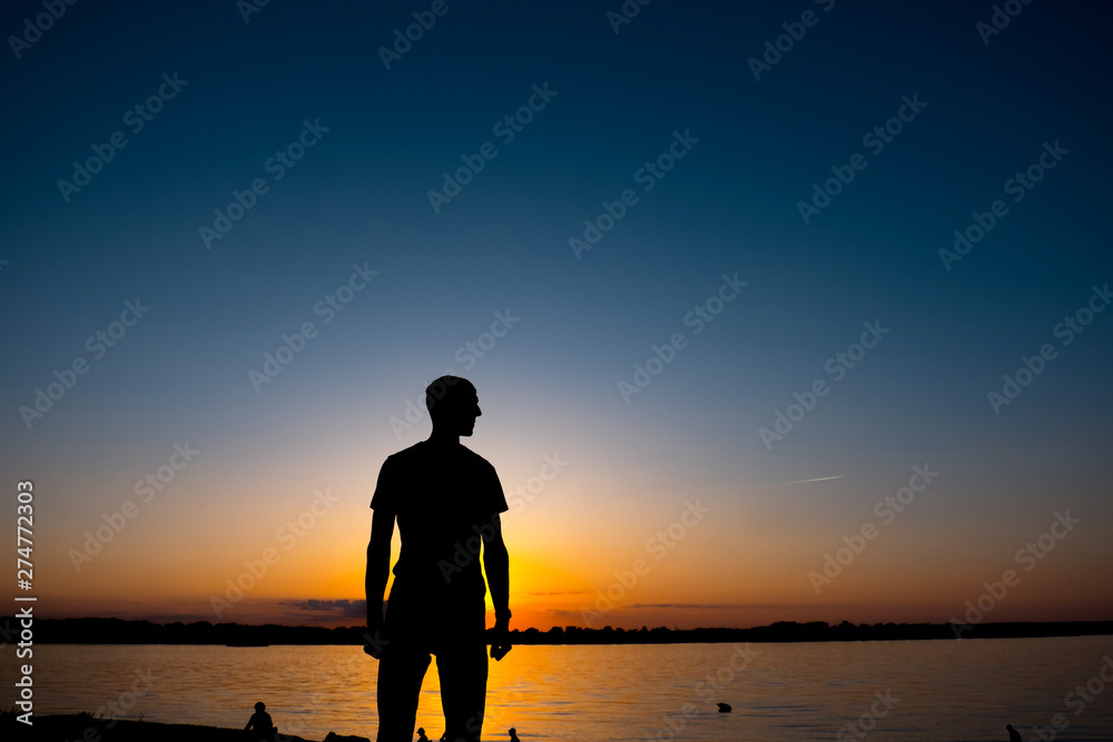 The young man is standing before the river bank with yesllow - blue Sunrise or sunset