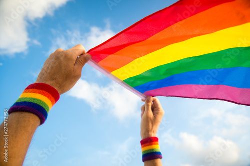 Hands with rainbow color wristbands waving gay pride flag backlit in the wind against a soft blue sky