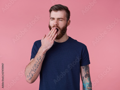 Photo of young handsome tired red bearded manman in blank t-shirt, wonts to sleep and yawns, stands over pink background with wide open mouth and eyes. photo