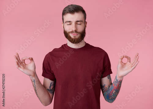 Portrait of young attractive red bearded man in blank t-shirt, looks peaceful and calm, smiles, stands over pink background with closed eyes and showing om gesture.