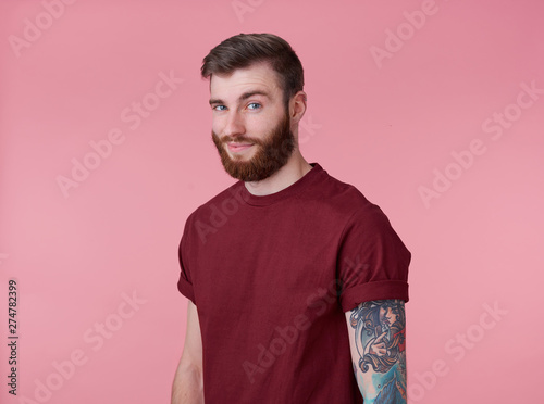 Portrait of young handsome happy red bearded tattooed man in red t-shirt, looks cheerful and broadly smiles, looks away, stands over pink background.