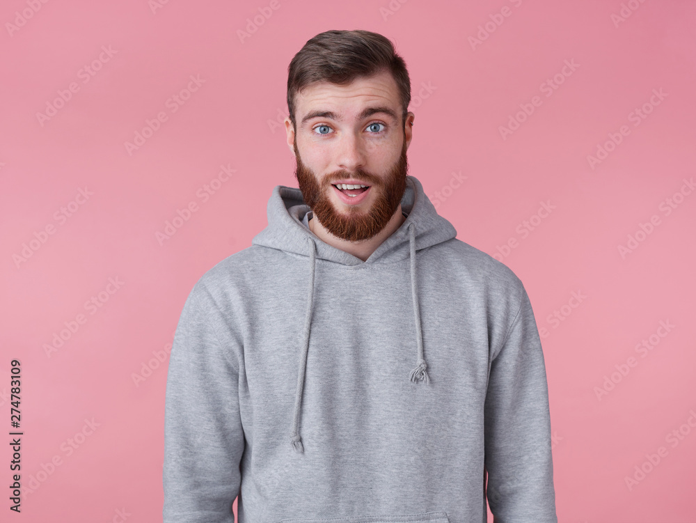 Hah, really? Young handsome red bearded man in gray hoodie looks surprised, stands over pink background looks at the camera with amazed.