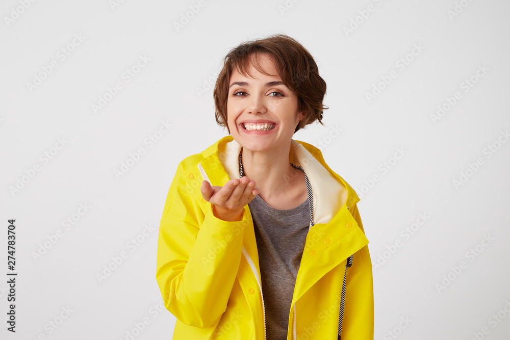Portrait of young happy cute short haired girl wears in yellow rain coat, broadly smiles and pointing palms at the camera, like holding something small in the palm, stands over white wall. Portrait of young happy cute short haired girl wears in yellow rain coat, broadly smiles and pointing palms at the camera, like holding something small in the palm, stands over white wall.