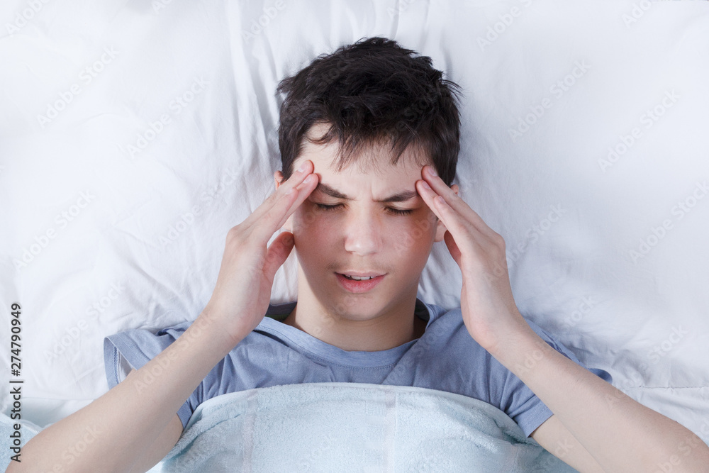 the severe headache, the teenager, the boy keeps hands on the head in a bed the severe headache, the teenager, the boy keeps hands on the head in a bed