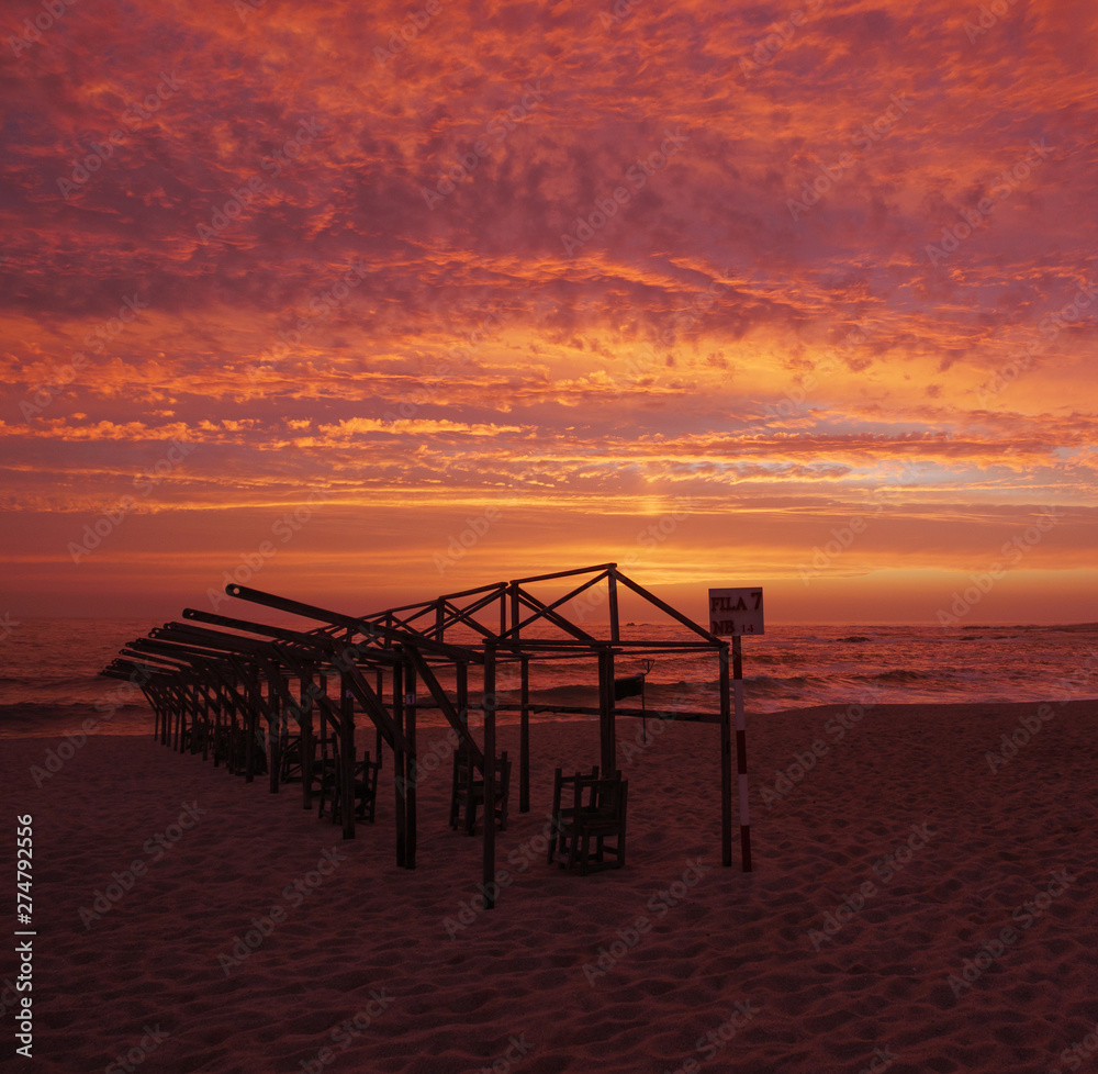 Beach hut frames silhouetted against vivid red sunset sky with dramatic clouds, on beach in Portugal