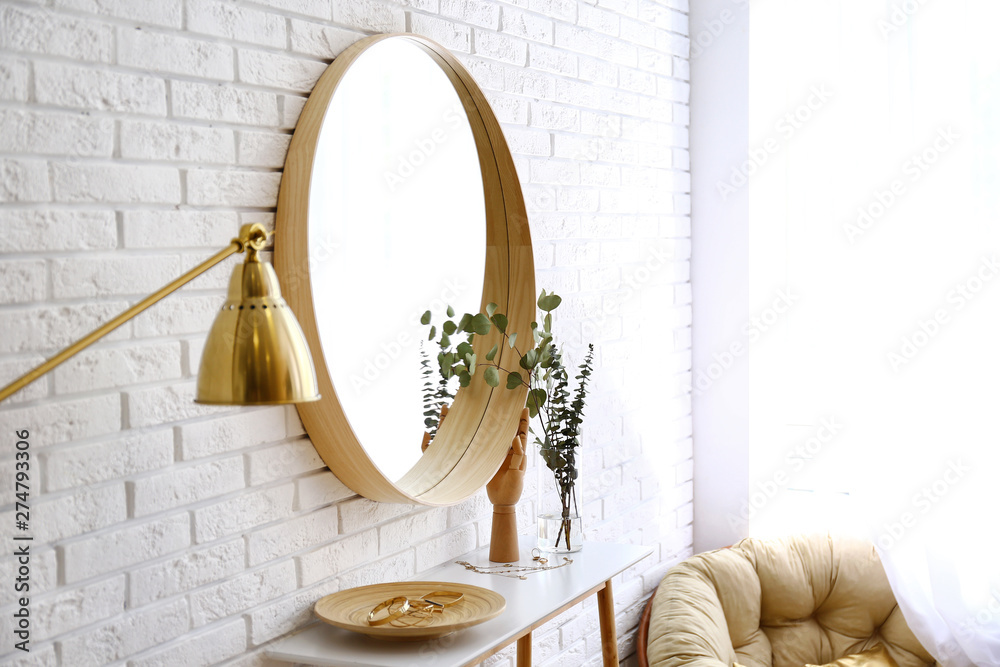 Big round mirror, table with jewelry and decor near brick wall in