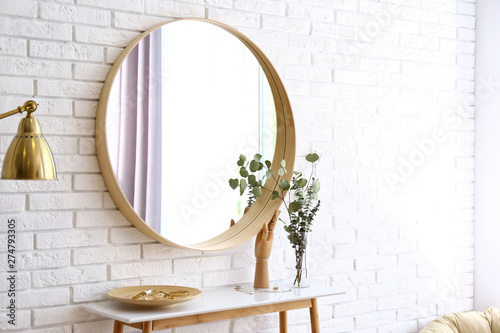 Big round mirror, table with jewelry and decor near brick wall in hallway interior photo