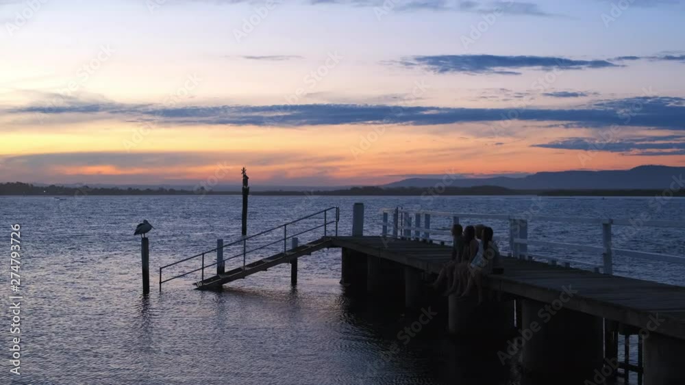 Handheld, tracking shot of people, sitting on a wooden dock and a Pelican, on a pole, dawn, at Culburra Beach, in New South Wales, Australia Handheld, tracking shot of people, sitting on a wooden dock and a Pelican, on a pole, dawn, at Culburra Beach, in New South Wales, Australia