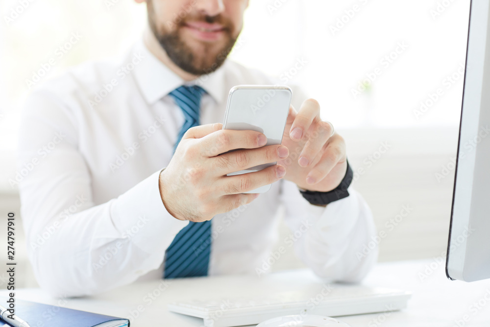 Close-up of content young businessman in white shirt sitting at desk with modern computer and viewing message on smartphone
