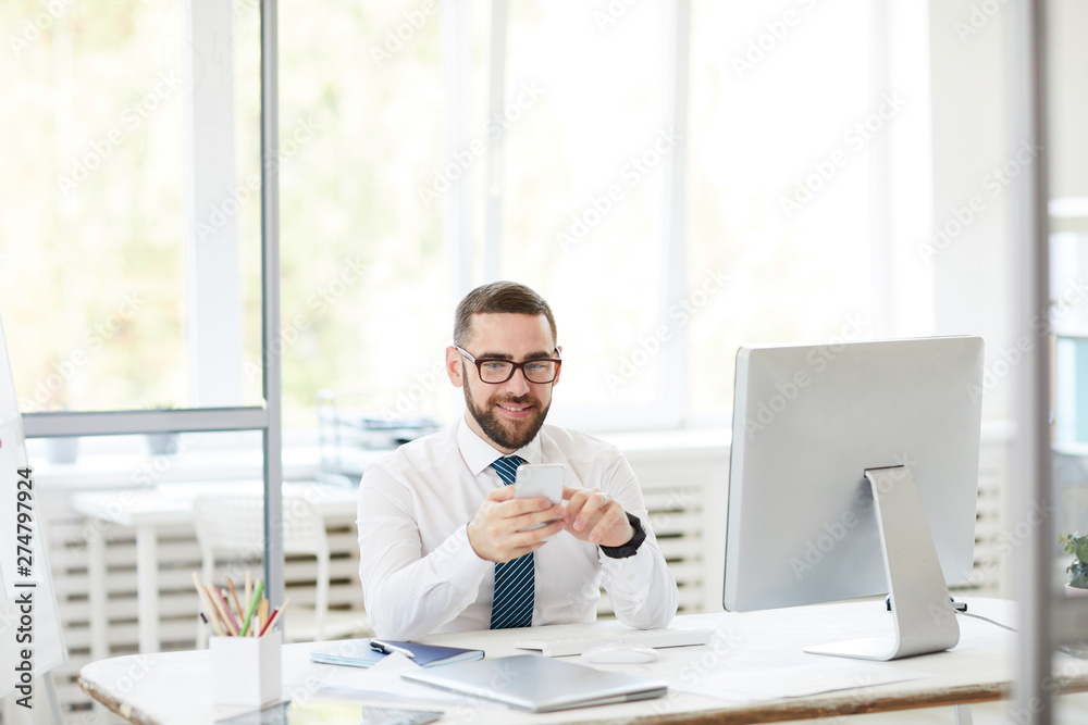 Smiling confident handsome young bearded manager in eyeglasses sitting at table and using smartphone while working in office Smiling confident handsome young bearded manager in eyeglasses sitting at table and using smartphone while working in office