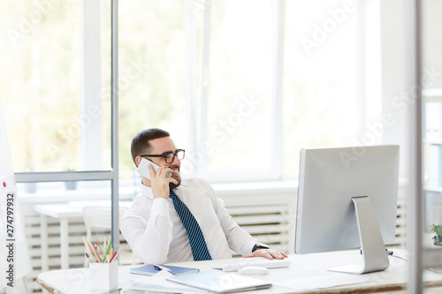 Cheerful handsome young bearded manager in eyeglasses leaning on table and calling colleague from office