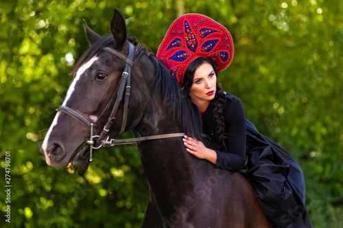 Gorgeous Model in Russian Style Kokoshnik Straddling On the Thoroughbred Horse. Posing Against Nature Background. photo