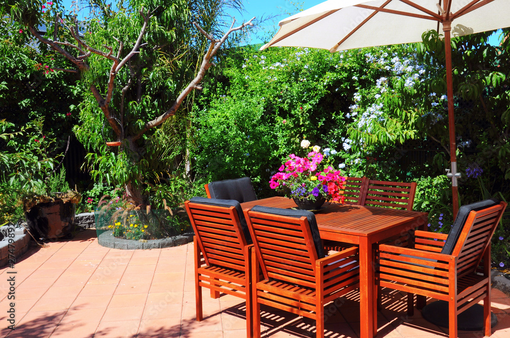 Fototapeta Beautiful and lush summertime Mediterranean style courtyard garden with wooden table and chairs and white market umbrella.