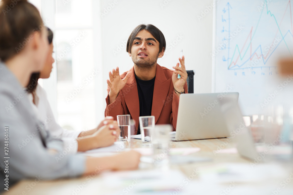 Portrait of young Middle-Eastern businessman talking to colleagues while discussing strategy during meeting in office, copy space Portrait of young Middle-Eastern businessman talking to colleagues while discussing strategy during meeting in office, copy space