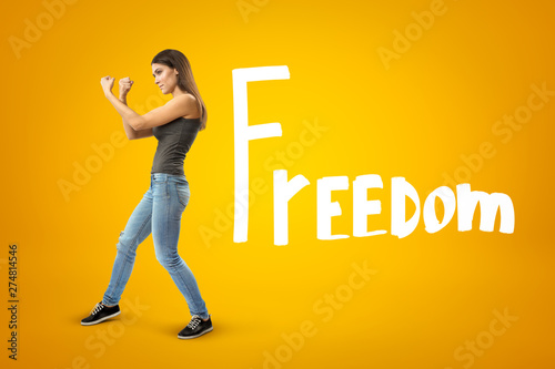 Young brunette girl wearing casual jeans and t-shirt showing double fist gesture with FREEDOM sign on yellow background
