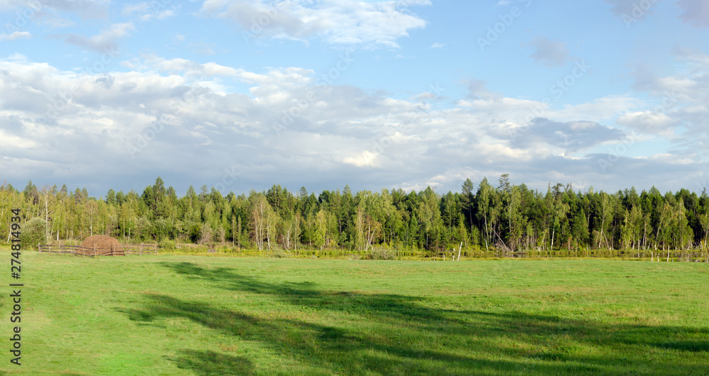 A haystack behind the fence stands on the edge of a green glade with shadows from large trees in the wild taiga of Yakutia.