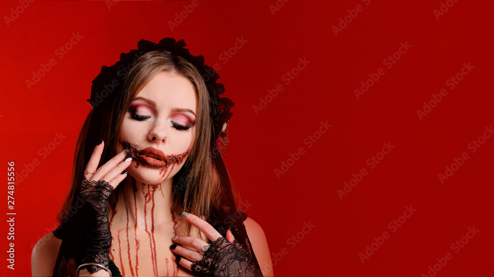 All Saints Day. Beautiful girl with terrible makeup for Halloween holds fingers of her hands near the bloody mouth on a red background. Copy space. Blank for poster or card design. All Saints Day. Beautiful girl with terrible makeup for Halloween holds fingers of her hands near the bloody mouth on a red background. Copy space. Blank for poster or card design.