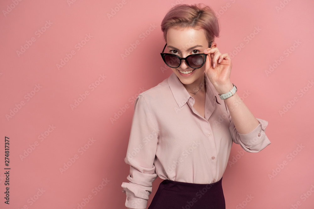 Pink-haired young woman looks through her sunglasses at the camera, pink background
