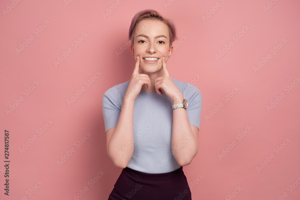 Pink-haired young woman looks at the camera pulling a smile on her face, pink background