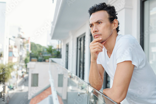 Pensive Vietnamese man rubbing chin and looking at city streets when standing on bacony of hotel room photo