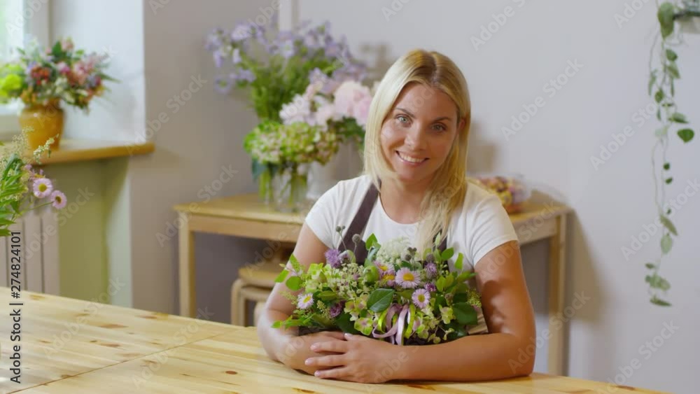Panning portrait shot of young blonde florist with bright delighted smile sitting at wooden table in flower shop and posing for camera, lovingly presenting her dainty floral arrangement Panning portrait shot of young blonde florist with bright delighted smile sitting at wooden table in flower shop and posing for camera, lovingly presenting her dainty floral arrangement