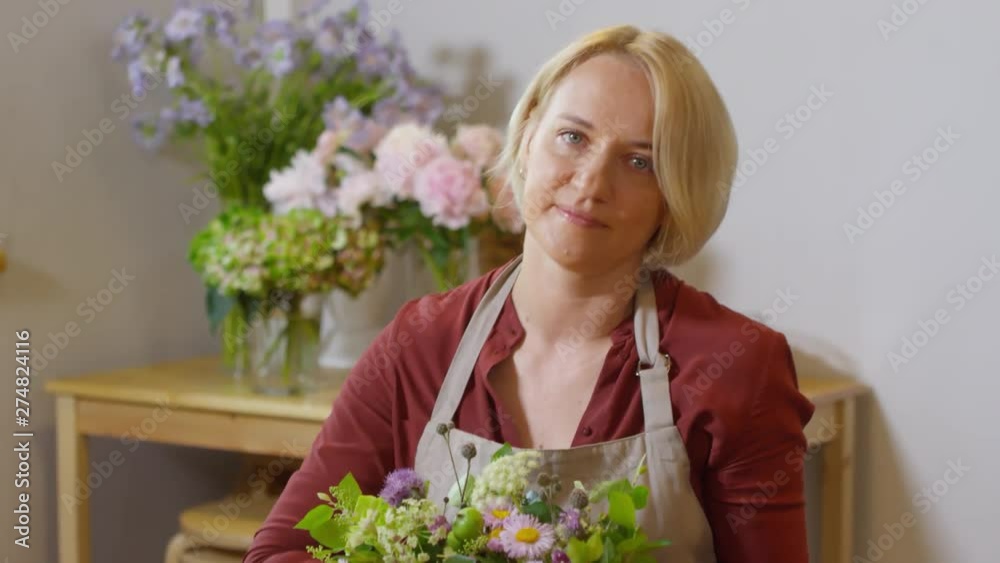 Tilting portrait shot of 30-something blonde floral designer at work in flower shop, posing for camera with modest smile and presenting her tasteful floral composition made with plain floral fillers Tilting portrait shot of 30-something blonde floral designer at work in flower shop, posing for camera with modest smile and presenting her tasteful floral composition made with plain floral fillers