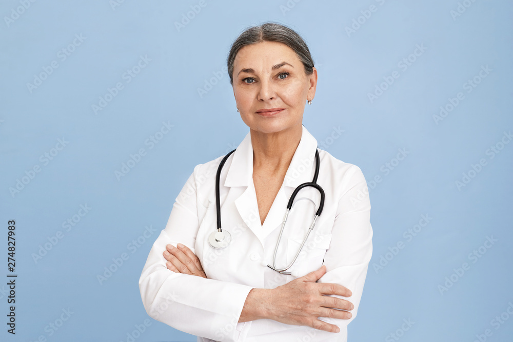Attractive confident sixty year old female surgeon wearing white uniform and stethoscope keeping arms folded on her chest posing isolated against blue wall background with copyspace for your content Attractive confident sixty year old female surgeon wearing white uniform and stethoscope keeping arms folded on her chest posing isolated against blue wall background with copyspace for your content