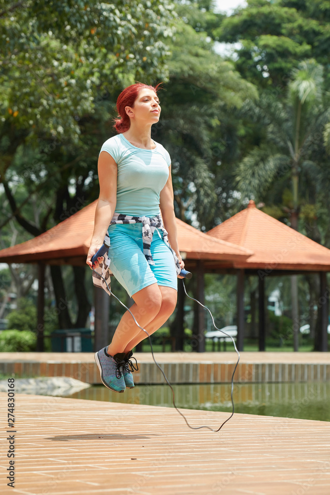 Sporty young woman jumping with skipping rope in summer park